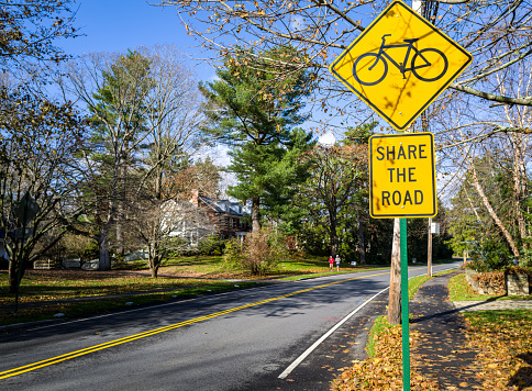 Suburban Boston town that encourages bicycles to share the road with cars.  Bright signage on double yellow lane road. Joggers are in the background.