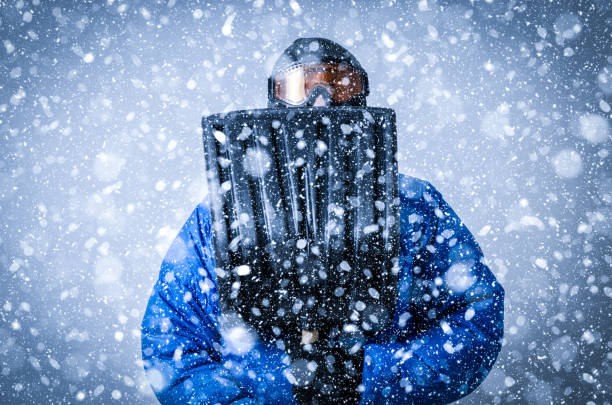 Man with goggles, tuque, winter coat hiding behind shovel during snowstorm