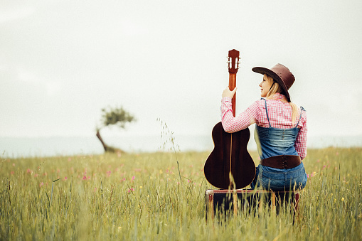 Beautiful blond cowgirl sitting in the meadow on her vintage suitcase with her the guitar. Creative color editing with added grain