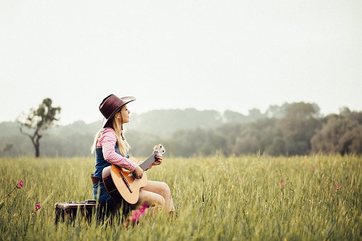 Blond cowgirl sitting in the meadow on her vintage suitcase playing the guitar. Creative color editing with added grain