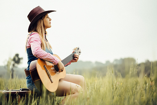 Portrait of a blond  cowgirl sitting on her vintage suitcase playing the guitar. Creative color editing with added grain