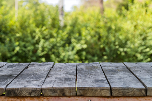 Grey wooden picnic table top at sunny summer day on blurred background of green bushes. Texture, background, poster, wallpaper