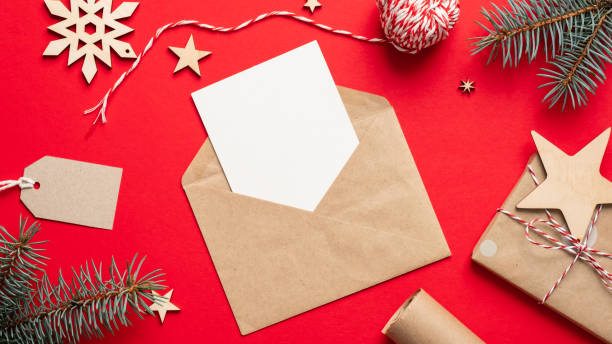 istock Vintage Christmas flat lay composition. Envelope, blank white card, gift box wrapped craft paper, Xmas wooden decorations, fir tree branches on red background. Christmas letter for Santa Claus concept 1285823759