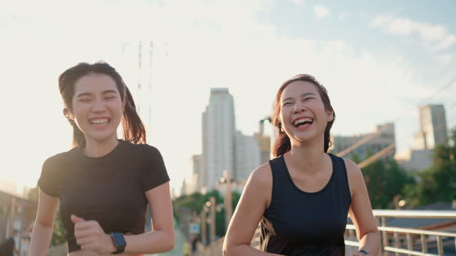 Two Asian woman in black sport cloth jogging and checking her smart watch at public park. They are smiling and are happy to enjoy this sunny day outside.