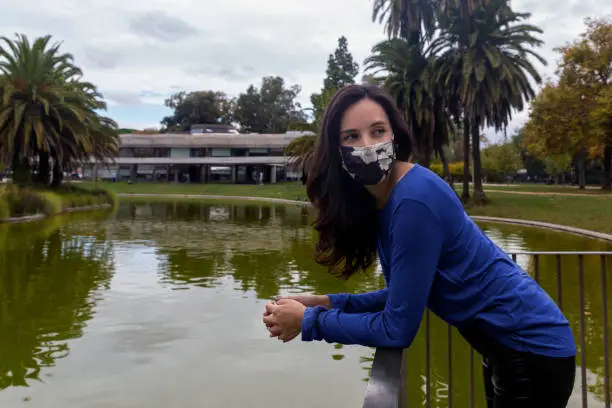 Young woman with long black hair, wearing a blue shirt at the park with social mask due to covid-19