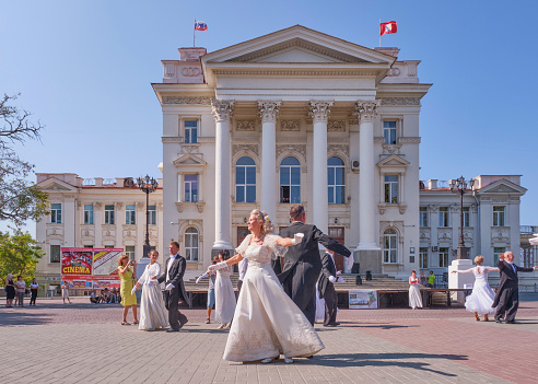 Sevastopol, Russia - September 29, 2020: Elderly couples dance a waltz over background of building of the Lunacharsky Drama Theater. Women in beautiful vintage white ballroom dresses, men in tuxedos.