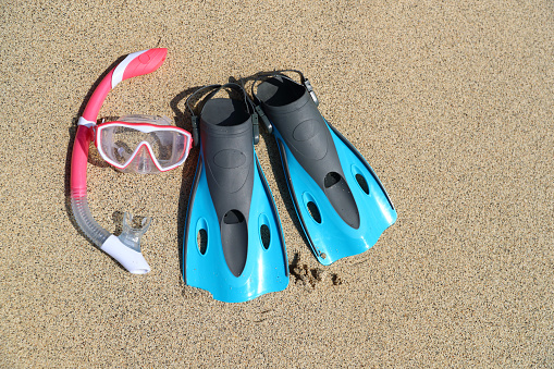 Beach vacation fun snorkel equipment with ocean waves splashing water. Scuba diving and snorkelling. Blue Flippers, pink mask, snorkel on sandy texture background. Objects lying on sand.