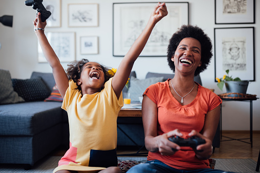 Single African American mother playing video games with her young daughter at home, competing and enjoying themselves, smiling
