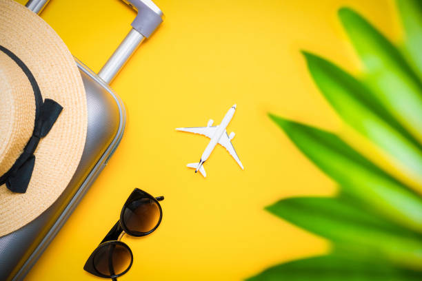 istock Summer background table. Suitcase, sunglasses with palm leaves and straw hat, white plane in travel composition on yellow background. Copy space, summer vacation and business travel concept. 1285852441