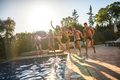 Front view of Spanish friends in 20s and 30s running and jumping together into swimming pool at party.