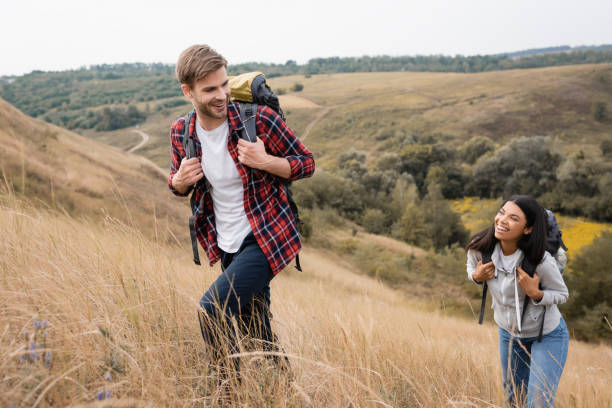 istock Smiling man with backpack walking near african american girlfriend on hill with grass 1285869347