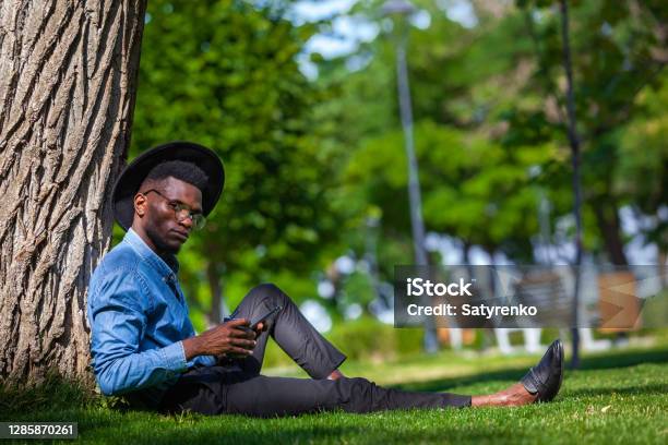 Portrait Of A Young Black Man In A Hat And Glasses Sitting Under Tree In The Park Holding A Cup Of Coffee In One Hand And A Mobile Phone In The Other Stock Photo - Download Image Now