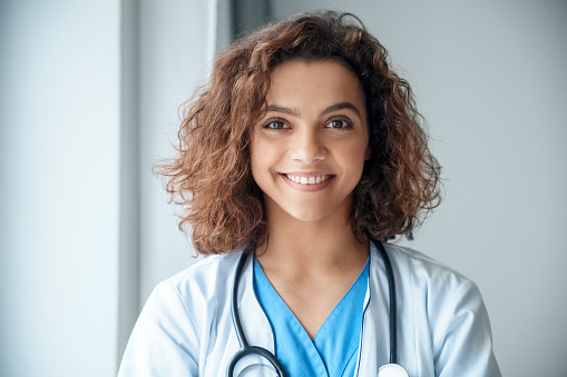 Close-up of happy young female doctor wear blue uniform, white medical coat, stethoscope and looking at camera in hospital office. Portrait of beautiful Hispanic female doctor, therapist, nurse.