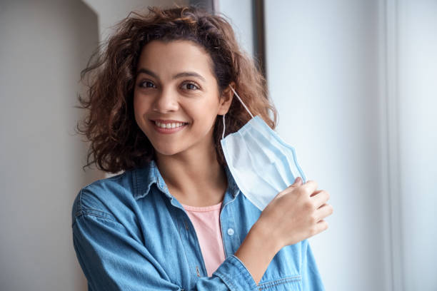 happy hispanic young woman takes off protective mask indoors. - removendo imagens e fotografias de stock