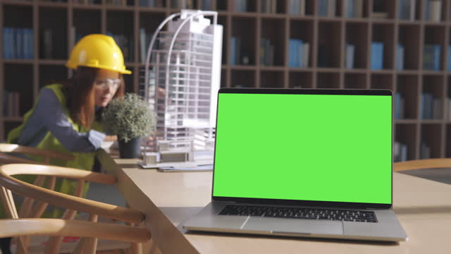 laptop computer with blank green screen on the table and female architects working on construction project of building