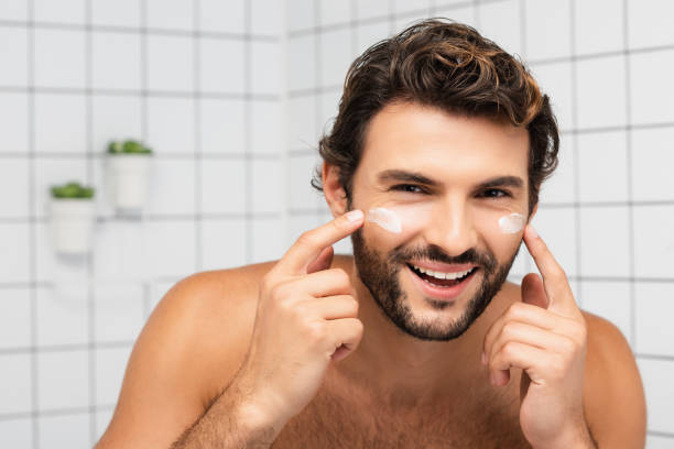 istock Cheerful shirtless man applying face cream and looking at camera in bathroom 1285882120