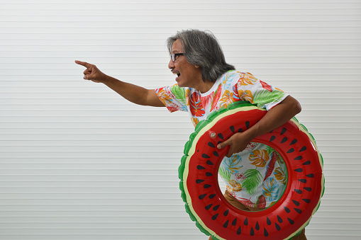 Happy elderly traveler asian man wearing summer shirt and eyeglasses holding watermelon inflatable ring pointing finger away running over white wall background, Business summer holiday concept