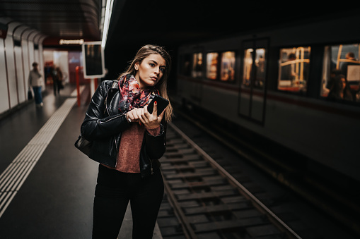 Young female commuter standing and texting in a subway while waiting for the train