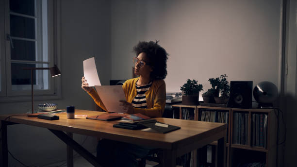 밤 늦게 집에서 일하기: 그녀의 홈 오피스에 앉아 문서를 읽는 젊은 아프리카 계 미국인 여성 - women black lighting equipment business 뉴스 사진 이미지