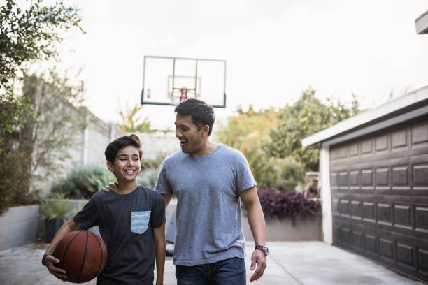 padre e hijo hispanos caminando de vuelta dentro después de jugar baloncesto al aire libre - bouncing ball family playing fotografías e imágenes de stock