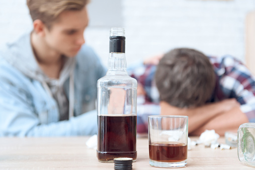 Drunk father is sitting at table with glass and bottles, drinking. His son is helpless to stop him.