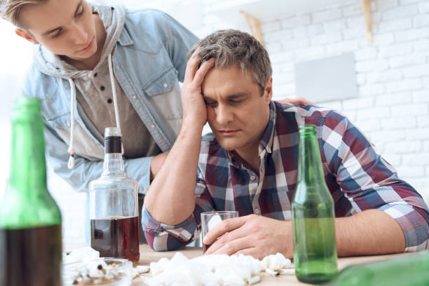 istock Drunk father is sitting at table with glass and bottles, drinking. 1285907889