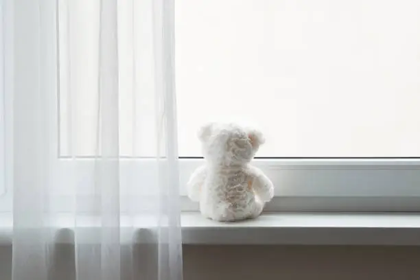 Photo of Sad white teddy bear alone sitting on window sill and looking out of home. Rear view. Waiting concept. Empty place for text quote or saying on glass.