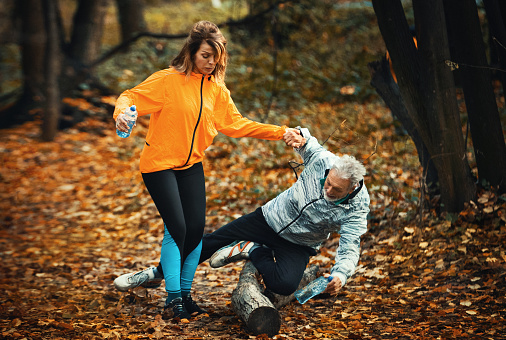 Closeup front view of a senior couple jogging in a forest and having fun. The next moment gentleman stumbles over tree trunk and falls.