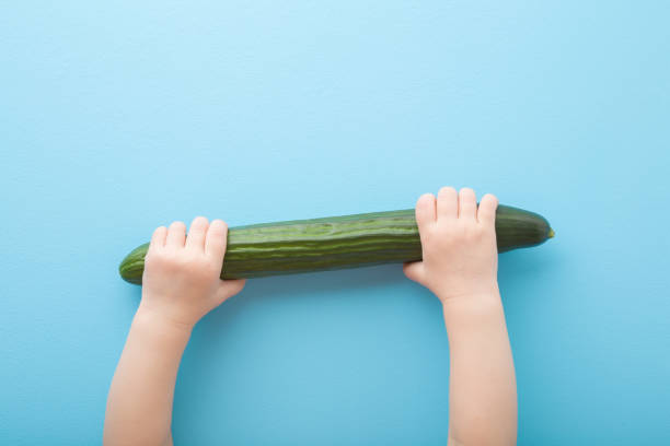 le mani della bambina che tengono il cetriolo lungo verde sullo sfondo del tavolo azzurro. colore pastello. verdure fresche. primo piano. colpo di punto di vista. vista dall'alto verso il basso. - pastel colored foto e immagini stock