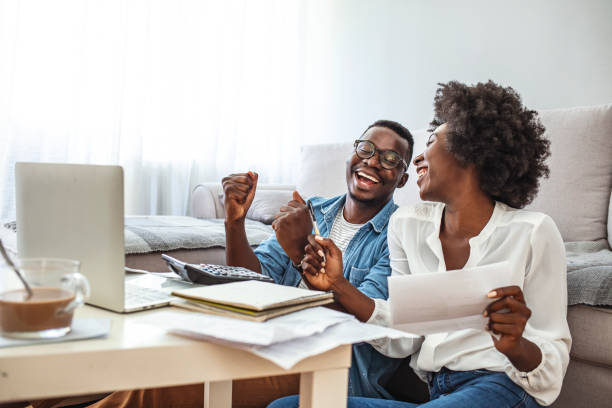 Happy couple with laptop spending time together at home. Family insurance concept. Positive black couple reading documentation at home, checking agreement details together. Young African Couple Sitting On Sofa Calculating Invoice bill legislation photos stock pictures, royalty-free photos & images
