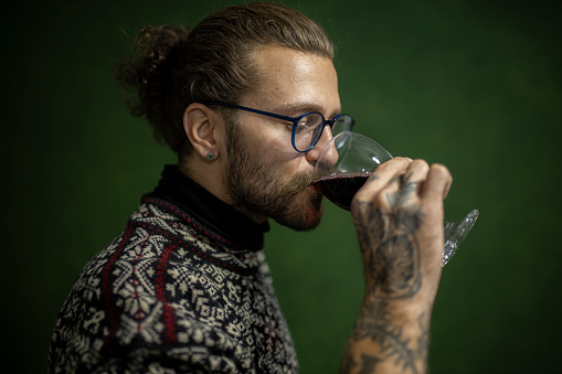 Young man with beard and eyeglasses drinking wine. He is holding glass of wine.