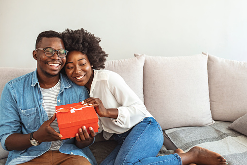 Beautiful young couple is celebrating at home. Handsome man is giving his girlfriend a gift box. Handsome man is giving his girlfriend a red gift box for birthday surprising in the living room