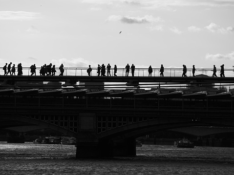 Peaceful, quiet image of some of the bridges over the River Thames. Image shows a silhouetted Blackfriars Rail Bridge and the Millennium Footbridge with people walking and enjoying the scenery. A dark, textured Thames is visible in the bottom half of the image and a cloud-spotted sky is overhead, with a single silhouetted bird near the centre.