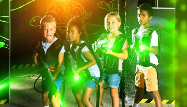 Portrait of multiracial group of smiling preteen kids with laser guns during lasertag game on dark arena