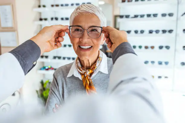 Smiling senior woman trying out new reading glasses at optician office. The doctor is putting on the glasses to see if it fits her.