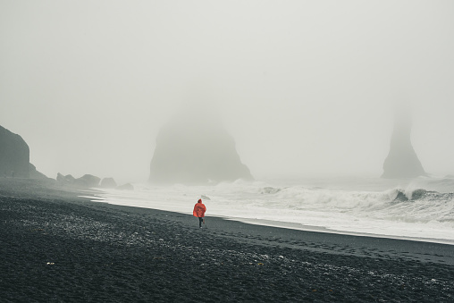 Famous black sand beach Vik in Iceland. Person in red raincoat running by the sea shore in the fog