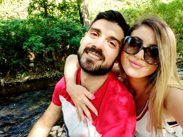 Young affectionate couple sitting near river and make selfie