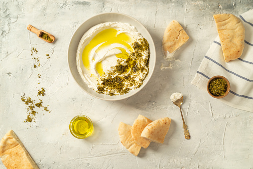 Popular middle eastern appetizer labneh or labaneh, soft white goat milk cheese with olive oil, hyssop or zaatar, served with pita bread on a grey table, top view, flat lay.
