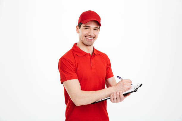 istock Content young delivery man 20s in red t-shirt and cap looking on camera while holding clipboard and writing down information, isolated over white background 1285940646