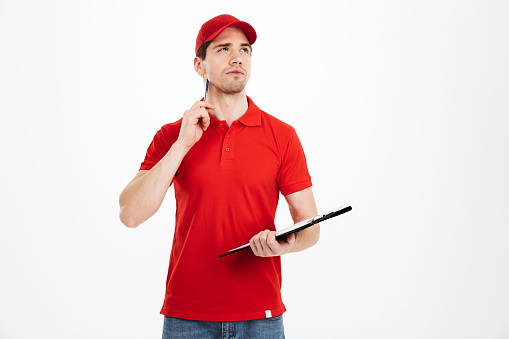Young courier man in red uniform looking upward with brooding view and holding clipboard isolated over white background