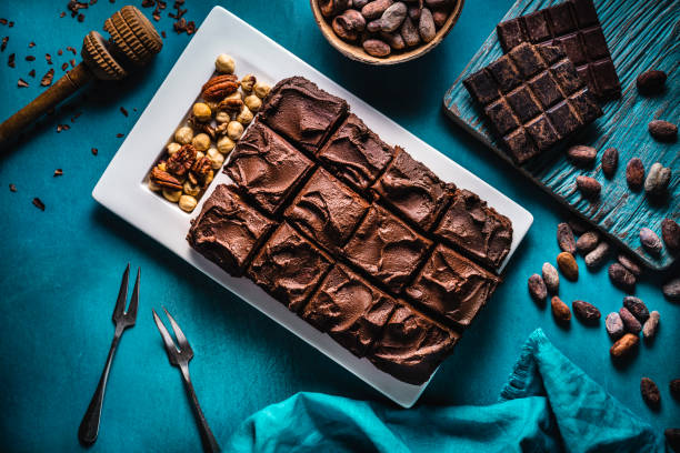 Brownie chocolate cake on turquoise background with nuts forks and linen napkin, cocoa beans and rustic chocolate bar