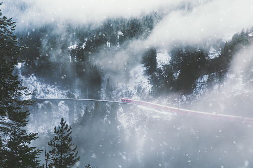 Scenic view of the red train riding through the pine forest, clouds and snowfall in the mountains in Switzerland