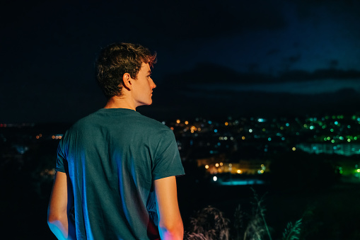 Rear view of a teenager man looking at the city lights at night. He is wearing a casual green t-shirt.