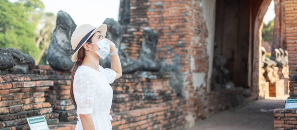 tourist woman in weißen kleid trägt chirurgische gesichtsmaske, schutz covid-19 pandemie während des besuchs im wat chaiwatthanaram tempel in ayutthaya. neues normal-, sicherheitsreise- und thailand-reisekonzept - wat chaiwattanaram stock-fotos und bilder