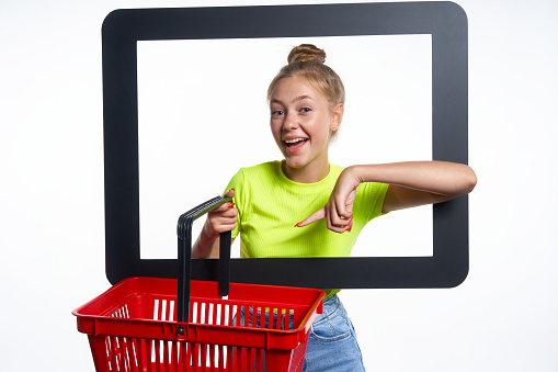 Online shopping concept. Trendy teen girl looking through digital screen holding empty shopping basket, pointing at it with surprise, isolated on light background