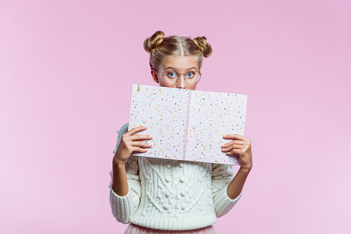 Portrait of happy teenager wearing white sweater and eyeglasses. High schools female student holding pink diary in hands. Studio shot on pink background.