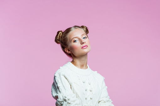 Portrait of confident teenager with hair buns wearing white sweater. High schools female student looking at camera. Studio shot on pink background.