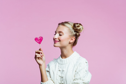 Portrait of teenager with hair buns wearing white sweater, holding heart shaped lollipop in hand. Studio shot on pink background.