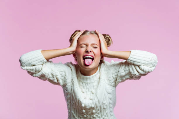 Cute teenege girl shouting with raised hands Portrait of surprised teenager with hair buns wearing white sweater. High schools female student shouting with raised hands with eyes closed and outstretched tongue. Studio shot on pink background. protruding stock pictures, royalty-free photos & images