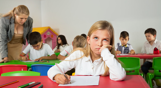 Portrait of upset girl sitting at lesson in schoolroom on background with pupils studying with teacher